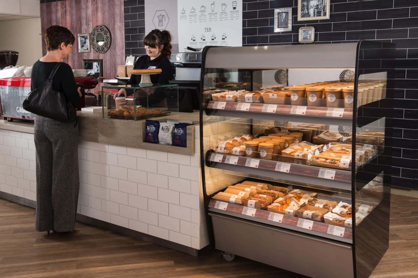 Café setting with a barista serving a customer, and a versatile food display filled with sandwiches and pastries